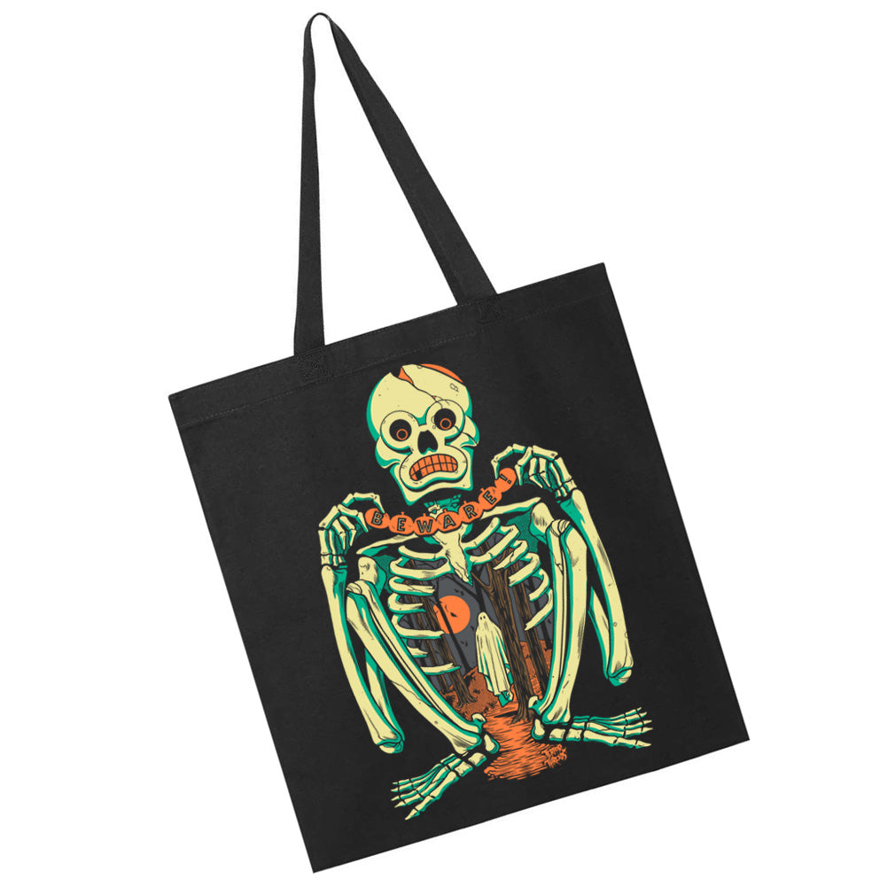 Terror Threads Original Beware black tote bag featuring a vintage Halloween skeleton holding a warning banner with a ghost, haunted graveyard, and orange moon.